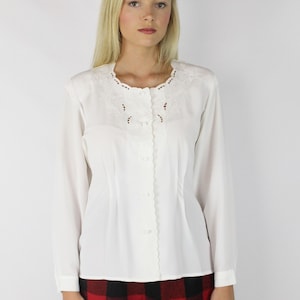 May include: A white long-sleeved blouse with a scalloped neckline and delicate floral embroidery. The blouse has a button-up front and a fitted silhouette.