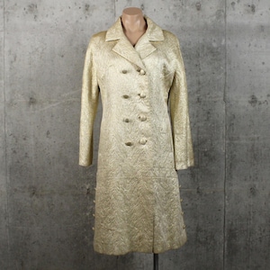 May include: A long-sleeved, double-breasted coat in a shimmering metallic gold fabric with a textured, swirling pattern. The coat features a notched collar, eight fabric-covered buttons arranged in two vertical rows, and a horizontal seam at the waist.