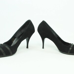 Op de afbeelding: Een paar zwarte su&egrave;de pumps met een spitse neus en dunne stiletto hakken. De schoenen hebben een zwart lint detail over de wreef.