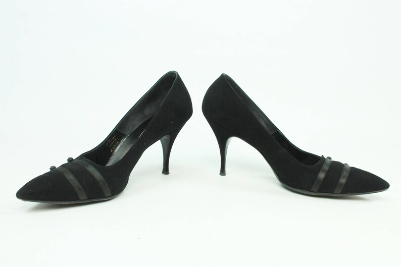 Op de afbeelding: Een paar zwarte su&egrave;de pumps met een spitse neus en dunne stiletto hakken. De schoenen hebben een zwart lint detail over de wreef.