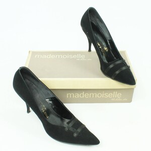 Op de afbeelding: Twee paar zwarte su&egrave;de pumps met een spitse neus en een dun zwart lintdetail. De schoenen staan op een doos met de tekst "mademoiselle" erop.