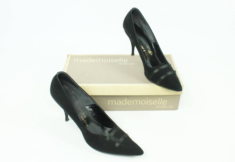 Op de afbeelding: Twee paar zwarte su&egrave;de pumps met een spitse neus en een dun zwart lintdetail. De schoenen staan op een doos met de tekst "mademoiselle" erop.