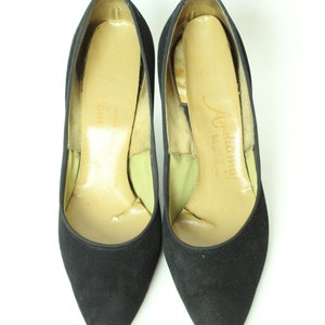 May include: A pair of black suede high heels with a pointed toe. The shoes have a light brown leather insole with the text "J. Adamy! Made in Italy" printed on the insole.