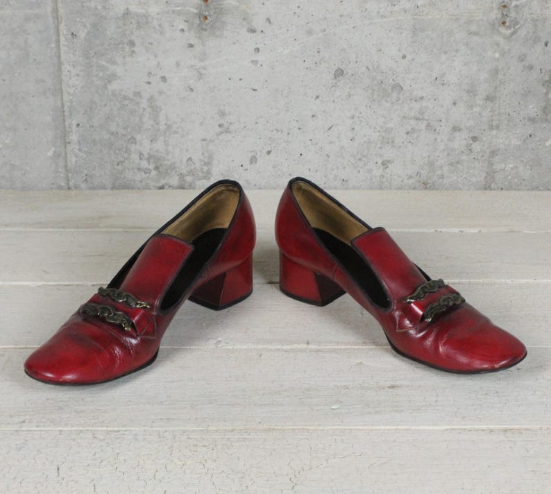 K&ouml;nnte beinhalten: Ein Paar dunkelrote Leder-Loafer mit niedrigem Blockabsatz und abgerundeter Zehenpartie. Jeder Schuh verf&uuml;gt &uuml;ber ein dunkles, metallisches Zierkettendetail auf dem Vorderblatt und ein schwarzes Innenfutter mit einer polierten Oberfl&auml;che.