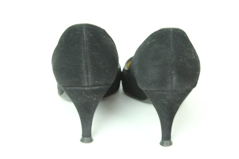 May include: A pair of black suede heeled shoes with a pointed toe. The shoes are shown from the back, with the heels facing the viewer.