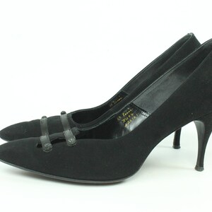 Op de afbeelding: Een paar zwarte su&egrave;de pumps met hoge hakken, een spitse neus en een decoratieve band over de wreef. De schoenen hebben een stilettohak en zijn in goede staat.