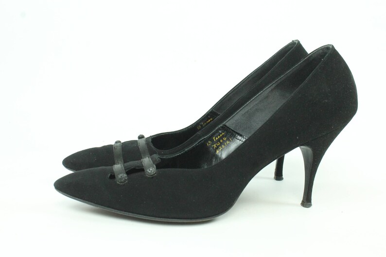 Op de afbeelding: Een paar zwarte su&egrave;de pumps met hoge hakken, een spitse neus en een decoratieve band over de wreef. De schoenen hebben een stilettohak en zijn in goede staat.