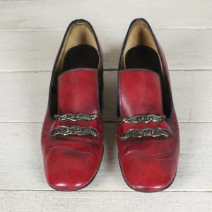 K&ouml;nnte beinhalten: Ein Paar rote Leder-Loafer mit abgerundeter Schuhspitze und zwei horizontalen, goldfarbenen, metallischen Zierkettenelementen &uuml;ber dem Vorderblatt. Die Schuhe haben ein dunkles Innenfutter und eine glatte, leicht abgenutzte Oberfl&auml;che.