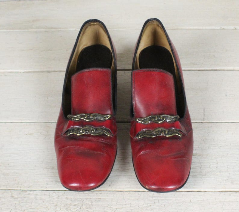 K&ouml;nnte beinhalten: Ein Paar rote Leder-Loafer mit abgerundeter Schuhspitze und zwei horizontalen, goldfarbenen, metallischen Zierkettenelementen &uuml;ber dem Vorderblatt. Die Schuhe haben ein dunkles Innenfutter und eine glatte, leicht abgenutzte Oberfl&auml;che.