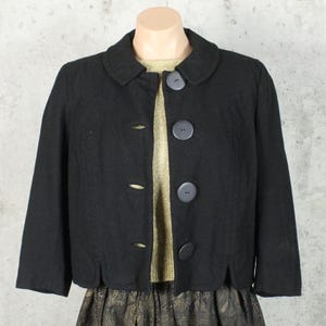 Puede incluir: Chaqueta negra de lana con cuello redondo y tres botones grandes de color gris oscuro. La chaqueta corta tiene mangas tres cuartos y se muestra en un maniquí. La chaqueta se combina con una blusa dorada y una falda estampada.