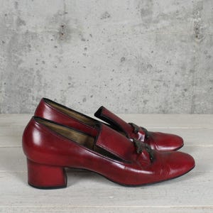 K&ouml;nnte beinhalten: Ein Paar dunkelrote Leder-Loafer mit Blockabsatz und einem dunklen, dekorativen Quastendetail auf dem Vorderblatt. Die Schuhe haben eine glatte Oberfl&auml;che mit schwarzem Besatz entlang des Schafts und eine abgerundete Zehenpartie im Slipper-Stil.