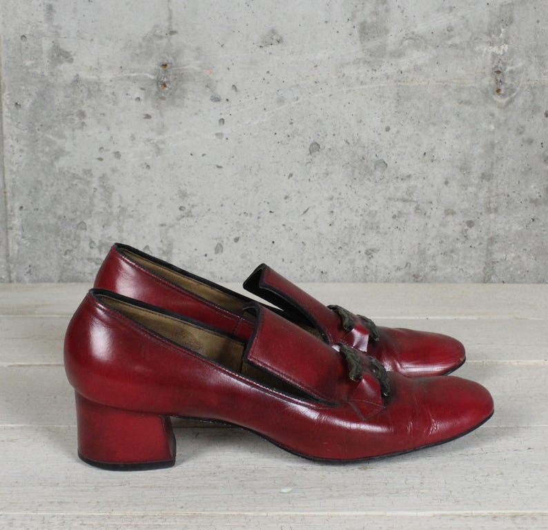 K&ouml;nnte beinhalten: Ein Paar dunkelrote Leder-Loafer mit Blockabsatz und einem dunklen, dekorativen Quastendetail auf dem Vorderblatt. Die Schuhe haben eine glatte Oberfl&auml;che mit schwarzem Besatz entlang des Schafts und eine abgerundete Zehenpartie im Slipper-Stil.