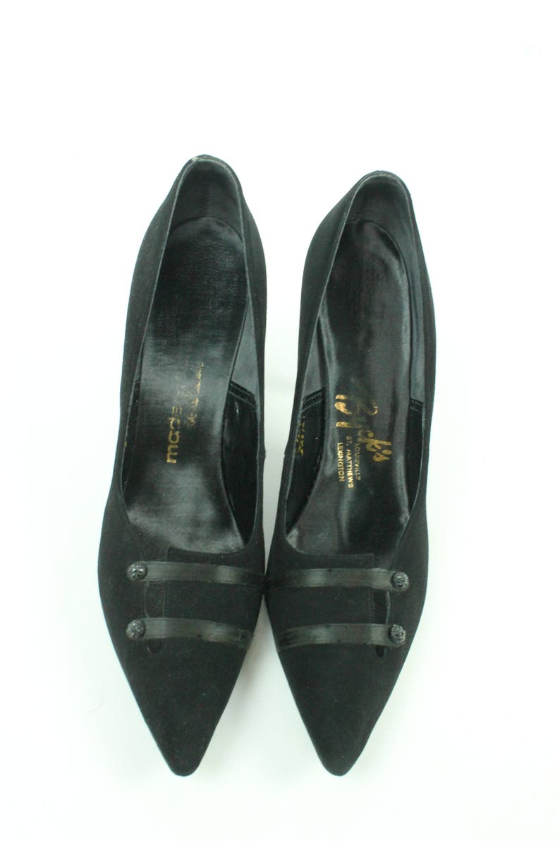 Op de afbeelding: Een paar zwarte su&egrave;de pumps met een spitse neus. De schoenen hebben een zwart satijnen lintdetail en een knoopaccent. De schoenen zijn gelabeld met "Slick's" en "Made in England".