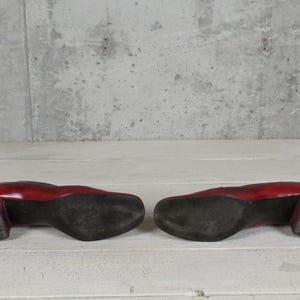 K&ouml;nnte beinhalten: Ein Paar rote Lederschuhe mit Blockabsatz, von unten betrachtet. Die Sohlen sind schwarz und weisen Gebrauchsspuren auf, mit einer glatten roten Oberfl&auml;che an den Seiten der Abs&auml;tze und der R&uuml;ckseite der Schuhe, mit quadratischer Zehenpartie.