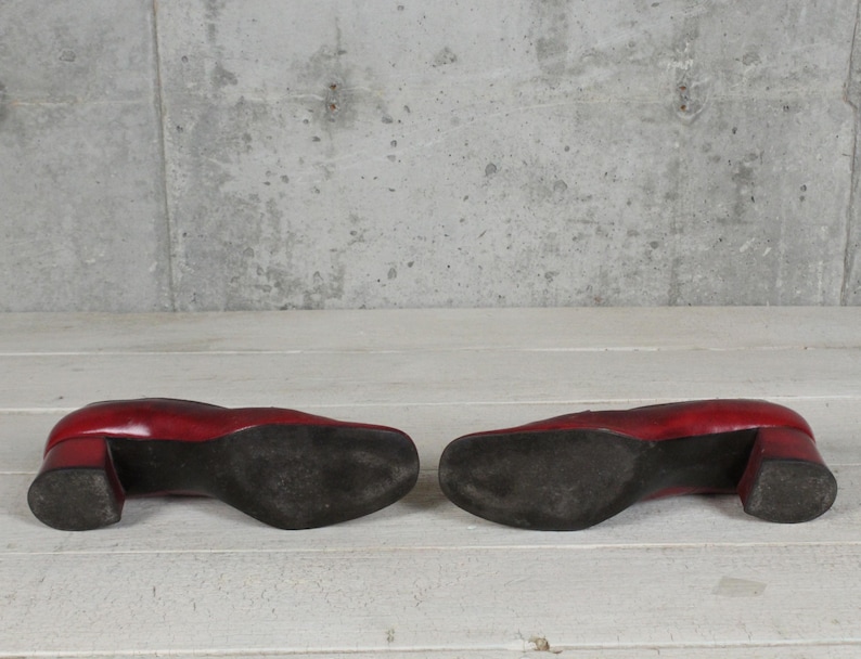 K&ouml;nnte beinhalten: Ein Paar rote Lederschuhe mit Blockabsatz, von unten betrachtet. Die Sohlen sind schwarz und weisen Gebrauchsspuren auf, mit einer glatten roten Oberfl&auml;che an den Seiten der Abs&auml;tze und der R&uuml;ckseite der Schuhe, mit quadratischer Zehenpartie.