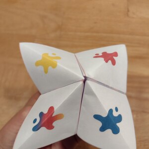 Könnte beinhalten: Ein weißer Papier-Orakel mit vier Klappen. Jede Klappe hat einen andersfarbigen Farbfleck: gelb, rot, blau und einen Farbverlauf von rot, orange und blau.