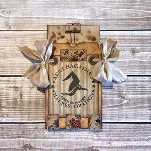 May include: A vintage-style sign with a witch's hat and the text "Aunt Hagatha's Hat Restoration Open 24 Hours" in a brown, white, and black color scheme.