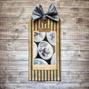 May include: A yellow and black striped fabric tag with a blue bow. The tag has a clear plastic window with a black and white image of floral designs on eggs. The text "Jiri Lomnicer" and "Kladno" is printed on the tag.