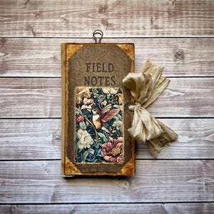May include: A rectangular journal with a brown cover featuring the text FIELD NOTES in black. The center displays a colorful illustration of a hummingbird among flowers. The journal has a metal ring at the top and a beige fabric ribbon tied on the side.