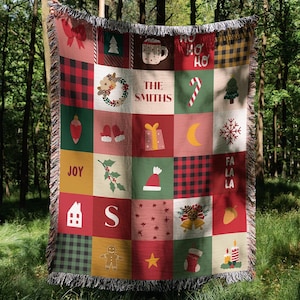 May include: A Christmas-themed throw blanket with a patchwork design. The blanket features squares with festive images like a Christmas tree, a snowflake, and a stocking. The blanket has a red, green, and gold color scheme and includes the text "The Smiths" and "Joy".