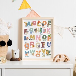 May include: A framed alphabet art print with colorful letters and animal illustrations. The print is displayed on a white surface with a woven basket, a wooden toy car, and a decorative bear-shaped cup. A plush bear head and decorative bunting are also visible.