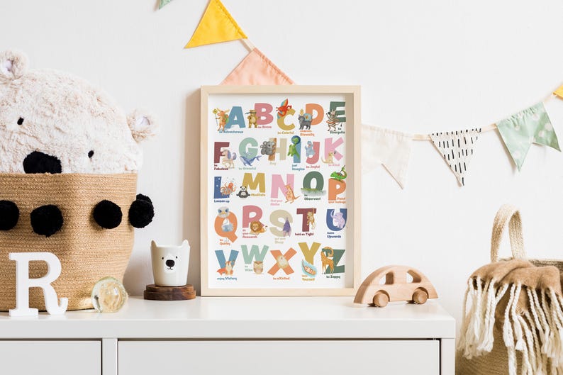 May include: A framed alphabet art print with colorful letters and animal illustrations. The print is displayed on a white surface with a woven basket, a wooden toy car, and a decorative bear-shaped cup. A plush bear head and decorative bunting are also visible.