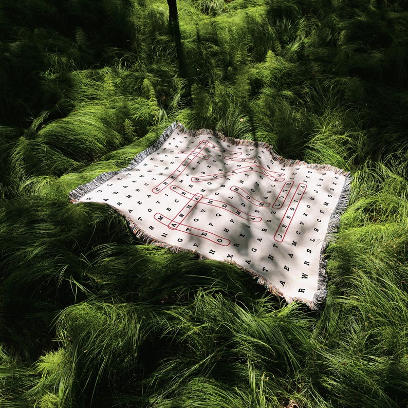 May include: A white woven blanket with a word search pattern printed on it. The blanket is laid out on a bed of green grass.