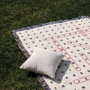 May include: A white woven throw with a red and black word search pattern. The throw has fringe on all sides. A white cushion is on top of the throw.