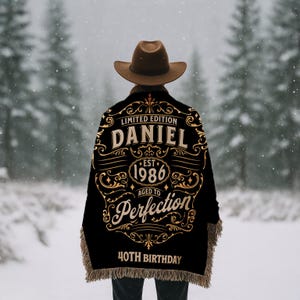 May include: A black throw blanket with gold lettering and fringe. The blanket reads "LIMITED EDITION DANIEL EST 1986 AGED TO PERFECTION 40TH BIRTHDAY". A person wearing a brown cowboy hat is wearing the blanket in a snowy forest.
