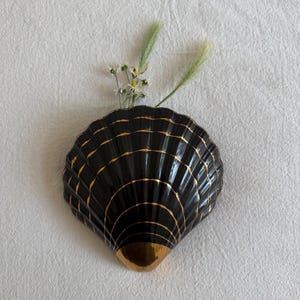 May include: A black ceramic wall vase shaped like a scallop shell, featuring thin gold horizontal lines along its ridges and a solid gold tip at the base. Small white daisy-like flowers and green grass stems are tucked inside the shell opening.