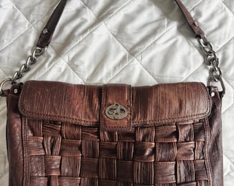 May include: A brown leather handbag with a woven pattern. The bag features a flap closure with a metal clasp and a leather strap with silver chain detailing. The bag is set against a white background.