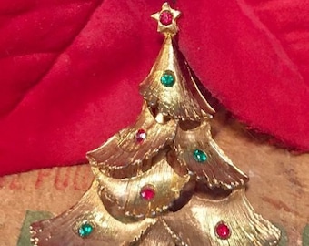 Puede incluir: Un broche de árbol de Navidad de tono dorado con diamantes de imitación rojos y verdes. El árbol está hecho de metal y tiene una estrella en la parte superior.