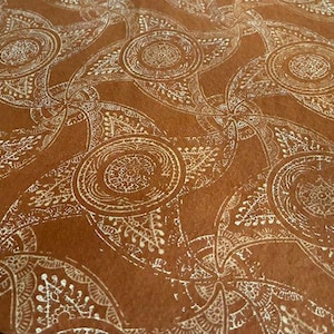 Könnte beinhalten: Nahaufnahme eines braunen Textils mit einem sich wiederholenden weißen Paisley-Muster. Das Design zeigt kreisförmige und tropfenförmige Formen mit aufwendigen Details und erzeugt eine strukturierte, dekorative Oberfläche.