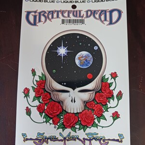 May include: A white sticker with a black and white illustration of a skull with a tear, surrounded by red roses. The skull has a black background with a white star and a blue and green planet. The text "GRATEFUL DEAD" is in blue and red letters above the skull. The text "SPACE YOUR FACE" is in purple and yellow letters below the skull.