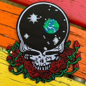 Puede incluir: Parche bordado con una calavera con un fondo negro que representa el espacio y un planeta. La calavera está rodeada de rosas rojas con hojas verdes. El parche está sobre una superficie de madera colorida.