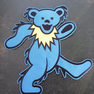 Può includere: Una toppa a forma di orso blu con colletto giallo e contorni neri. L'orso è in posa da ballo con le braccia e le gambe divaricate. La toppa è probabilmente per abbigliamento o accessori.