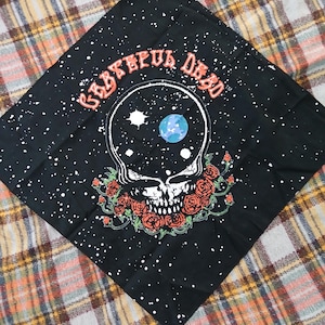 May include: Black bandana featuring a skull design with roses, a blue planet, and the words "Grateful Dead" in orange. The bandana has a white speckled pattern and is folded on a plaid surface.