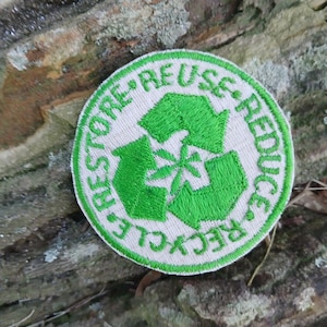 Puede incluir: Un parche bordado circular con fondo beige claro y un símbolo de reciclaje verde brillante en el centro. El borde contiene el texto bordado en verde REUSE, REDUCE, RECYCLE y RESTORE dispuesto en un patrón circular alrededor.