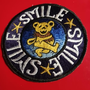Peut inclure: Un écusson rond, noir, brodé avec un contour blanc. L'écusson représente un ours de dessin animé avec un manteau de fourrure brun, un corps jaune et un fond bleu. Le texte "Smile Always Smile" est écrit en cercle autour de l'ours, avec des étoiles entre les mots.