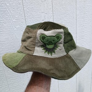 Op de afbeelding: Een patchwork bucket hat van corduroy stof in diverse tinten olijfgroen, saliegroen en beige. Het voorpaneel heeft een lichtgekleurde corduroy patch met een geborduurd groen berengezicht en een stervormige kraag in donkergroen en zwart.