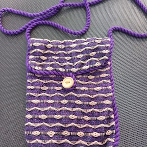 May include: A small purple and white striped fabric bag with a purple rope strap and a wooden button closure.