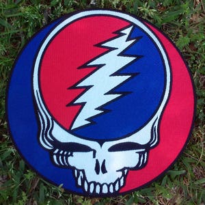 May include: A round patch featuring the iconic Grateful Dead 'Steal Your Face' skull logo. The design includes a red and blue background with a white lightning bolt and skull. The patch is set against a backdrop of green grass.