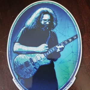 May include: A blue and white oval sticker featuring a black and white image of Jerry Garcia playing a guitar. The sticker has the text "© 2018 Jerry Garcia Family LLC under exclusive license to JGF Rights Holding, LLC." at the bottom.