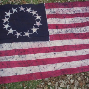 May include: A batik American flag with 13 red and white stripes and a blue canton featuring a circle of 13 white stars. The flag is made of a cotton fabric and has a faded, vintage look.