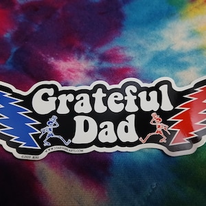 May include: A sticker with the words "Grateful Dad" in white, with a black border. The design includes blue and red stylized trees, and running skeleton figures. The background is a tie-dye pattern in shades of blue, purple, and red.