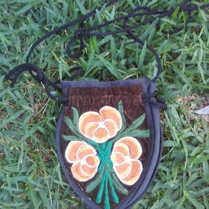 Könnte beinhalten: Eine kleine braune Samt-Zugschnur-Tasche mit einem schwarzen Band. Die Tasche hat ein gesticktes Design aus drei orangefarbenen Blumen mit grünen Blättern.