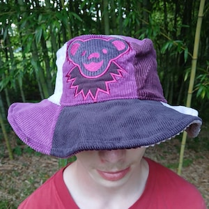 Könnte beinhalten: Ein Patchwork-Eimerhut aus lila, weißen und grauen Cordfarben. Der Hut hat ein gesticktes Bär-Design in Lila und Rosa. Der Rand ist ein dunklerer Lilaton. Der Hut ist ein lässiges Accessoire.