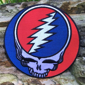 Può includere: Una toppa rotonda ricamata con l'iconico logo dei Grateful Dead, Steal Your Face. Il logo è una combinazione di un teschio e un fulmine, con uno schema di colori rosso e blu.