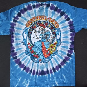 May include: A blue tie-dye t-shirt with a circular design featuring a skeleton in a blue robe, surrounded by roses and mushrooms. The text "Grateful Dead" is written in a circular arc above the skeleton.