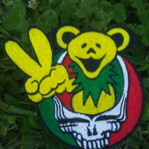 May include: Embroidered patch with a yellow bear peace sign, and a red, green, and white skull design. The patch is set against a background of green grass and clover.